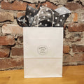White gift bag with polka dot bow against a brick wall